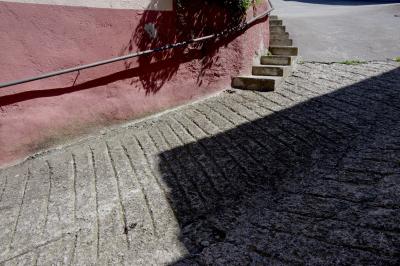 photograph “VI.2023 — Stairway to asphalt” par David Farreny — www.farreny.net — Espagne, Spain, España, Espanya, Catalogne, Catalonia, Cataluña, Catalunya, Lérida, Lleida, Alt Urgell, Ribera d'Urgellet, El Pla de Sant Tirs, rue, street, béton, concrete, asphalte, asphalt, bitume, blacktop, marches, steps, escalier, stairs, staircase, rampe, railing, métal, metal, mur, wall, virage, turn, angle, ombre, shadow, lignes, lines, rayures, grooves, plante, plant