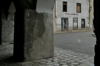 photograph “VI.2021” par David Farreny — www.farreny.net — France, Occitanie, Ariège, Tarascon-sur-Ariège, arcades, arches, archs, vieux, old, pavés, cobblestones, pierre, stone, boulangerie, bakery, fermée, closed down, magasin, shop, store, décadence, decay, crise, crisis, place, square, enseigne, sign, caractères, characters, lettres, letters, lampe, lamp, bitume, blacktop, asphalte, asphalt, misère, pauvreté, poverty, portes, doors, fenêtres, windows, ruine, ruin, câbles, cables, fils, wires, gouttières, drainpipes, trottoir, pavement, bâtiments, buildings