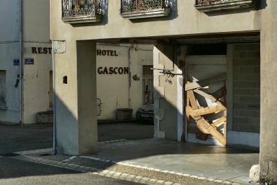 photograph “IV.2025 — Closedown” par David Farreny — www.farreny.net — France, Occitanie, Gascogne, Gascony, Lomagne, Tarn-et-Garonne, Lavit, Lavit-de-Lomagne, village, rue, street, bâtiment, building, maison, house, restaurant, hôtel, hotel, Le Gascon, fermé, closed down, porte, door, barricaded, barricadée, planches, planks, bois, wood, angle, coin, corner, balcons, balconies, enseigne, sign, ombre, shadow, plaques, plates, rue Matabiau, rue de Puygaillard, gouttières, drainpipes, métal, metal, store, parpaings, breeze-blocks, pierre, stone, voiture, car, fenêtres, windows, grille, railing, ciment, cement