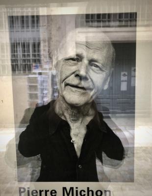 photograph “III.2018 — Self-portrait as a great double amputee writer” par David Farreny — www.farreny.net — France, Occitanie, Gascogne, Gascony, Gers, Auch, librairie, bookshop, vitrine, window, baie, bay, vitre, verre, glass, affiche, poster, Pierre Michon, écrivain, writer, homme, man, sourire, smile, photographie, photograph, noir et blanc, black and white, portrait, cadre, frame, manchot, double amputee, bras, arms, reflet, reflection, autoportrait, selfportrait, rue, street, pierre, stone, porte, door, grilles, railings