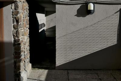 photograph “I.2023 — Faces of home, 163” par David Farreny — www.farreny.net — Andorre, Andorra, La Massana, immeuble, bâtiment, building, mur, wall, béton, concrete, pierre, stone, lampe, lamp, tuyaux, pipes, câbles, cables, fils, wires, grillage, fencing, ombre, shadow, passage, dalles, slabs, domicile, home
