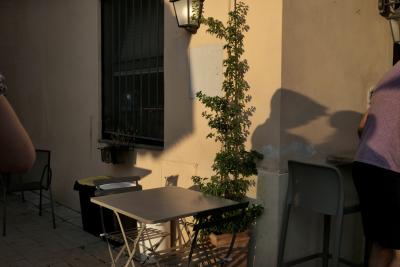 photograph “VIII.2022” par David Farreny — www.farreny.net — Italie, Italia, Italy, Toscane, Toscana, Tuscany, Arezzo, Cavriglia, été, summer, soir, evening, restaurant, terrasse, terrace, bâtiment, building, table, chaises, chairs, plante, plant, pot, fenêtre, window, lampe, lamp, coin, corner, angle, ombres, shadows, jeunes femmes, young women, coude, elbow, bras, arm, dos, back, souvenir, Li*, Lu*