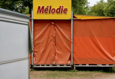 photograph “VI.2011” par David Farreny — www.farreny.net — France, Occitanie, Aveyron, Rouergue, Rodez, jardin, garden, Foirail, fête foraine, funfair, manège, ride, enseigne, sign, Mélodie, bâche, rideau, sheet, curtain, plastique, plastic, orange, jaune, yellow, arbres, trees, herbe, grass
