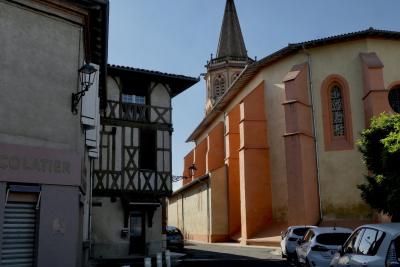 photograph “IX.2024 — Temple” par David Farreny — www.farreny.net — France, Occitanie, Haute-Garonne, Rieumes, église, church, temple, clocher, bell tower, rue, street, maisons, houses, magasin, shop, store, enseigne, sign, chocolatier, métal, metal, lampes, lamps, arbre, tree, voitures, cars, colombages, half-timbering, bois, wood, fenêtres, windows, porte, door, soir, evening, vitrail, stained glass, tuiles, tiles