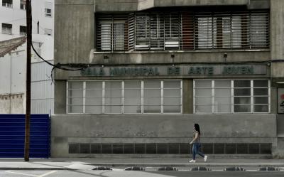 photograph “III.2023 — Arte joven” par David Farreny — www.farreny.net — Espagne, España, Spain, Aragon, Aragón, Saragosse, Zaragoza, Caesaraugusta, ville, city, rue, street, immeuble, bâtiment, building, béton, concrete, vieux, old, laid, ugly, baie, baie vitrée, bay window, fenêtres, windows, enseigne, sign, Sala municipal de arte joven, Salle municipale d'art jeune, Municipal hall of young art, poteau, post, pole, bois, wood, câbles, cables, fils, wires, palissade, palisade, clôture, fence, métal, metal, passante, passer-by, piétonne, pedestrian, femme, woman, jeune, young, brune, brunette, jeans, trottoir, pavement