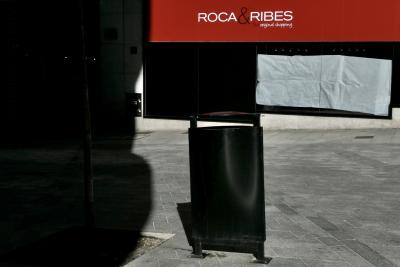 photograph “XI.2023 — Whatever” par David Farreny — www.farreny.net — Andorre, Andorra, Andorra la Vella, Andorre-la-Vieille, Andorra la Vieja, ville, city, rue, street, dallage, paving, poubelle, bin, trash, can, métal, metal, magasin, boutique, shop, store, enseigne, sign, Roca & Ribes, original shopping, vitrine, window, verre, vitre, glass, papier, paper, blanc, white, arbre, tree, ombre, shadow, pierre, stone, rouge, red