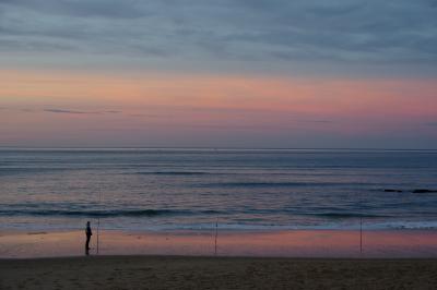 photograph “XII.2020 — Masked in front of infinity” par David Farreny — www.farreny.net — France, Aquitaine, Pyrénées-Atlantiques, Hendaye, Pays basque, Basque country, océan, Atlantique, Atlantic, ocean, mer, sea, eau, water, paysage, landscape, marin, seascape, vagues, waves, ciel, sky, soir, evening, hiver, winter, bleu, blue, rose, pink, pêcheur, fisher, fisherman, cannes à pêche, fishing rods, sable, sand, plage, beach, masque, mask, fucking COVID, homme, man, horizon, ligne, line, nuages, clouds, carte postale, postcard