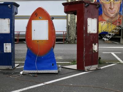 photograph “IV.2016” par David Farreny — www.farreny.net — France, Occitanie, Gascogne, Gers, Auch, fête foraine, funfair, fermée, closed, jeux, games, machines, métal, metal, bitume, asphalte, asphalt, blacktop, parking, car park, arbre, tree, platane, plane tree, câbles, cables, fils, wires, dos, back, marches, steps, escalier, stairs, illustration, peinture, painting, fresque, fresco, visage, face, homme, man, lunettes, glasses, yeux, eyes, bleus, blue, roues, roulettes, wheels, camion, truck