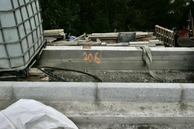 photograph “IX.2023 — 20.6” par David Farreny — www.farreny.net — Andorre, Andorra, Canillo, route, road, travaux, chantier, works, béton, concrete, trottoir, pavement, sol, soil, earth, pierre, stone, tuyau, pipe, serpent, snake, plastique, plastic, noir, black, nombre, number, peinture, paint, orange, 20.6, cailloux, pebbles, cuve, citerne, tank, métal, metal, bâche, sheet, lanière, tissu, strip, fabrication, palette, bois, wooden, pallet, planches, planks, arbres, trees, pelle, shovel
