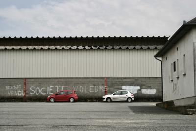 photograph “VIII.2011” par David Farreny — www.farreny.net — France, Occitanie, Aveyron, Rouergue, Réquista, bâtiment, building, entrepôt, warehouse, métal, metal, tôle ondulée, corrugated iron, parking, car park, bitume, blacktop, asphalte, asphalt, voitures, cars, mur, wall, paroi, parpaings, breeze-blocks, fenêtres, windows, nuages, clouds, graffiti, Bernat vendu, Société voleurs, Bernat crook, Société thieves