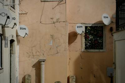 photograph “II.2020” par David Farreny — www.farreny.net — Portugal, Lisbonne, Lisboa, Lisbon, ville, city, capitale, capital, rue, street, paraboles, satellite, dishes, Zon, mur, wall, porte, door, fenêtre, window, fleurs, flowers, fils, wires, câbles, cables, colonne, column, panneau, sign, travessa do Chão da Feira, pierre, stone, profusion