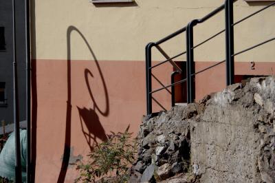 photograph “IX.2021 — Faces of home, 124” par David Farreny — www.farreny.net — Andorre, Andorra, Escaldes-Engordany, ville, city, immeuble, bâtiment, building, béton, concrete, mur, wall, automne, autumn, fall, gouttière, drainpipe, métal, metal, poteau, post, pole, lampadaire, lamp, ombre, shadow, sac, bag, plastique, plastic, rambarde, guardrail, pente, slope, pierre, stone, végétation, vegetation, plante, plant, fenêtres, windows, porte, door, numéro, number, 2, two, deux, jaune, yellow, rouge, red, domicile, home, boucle, loop