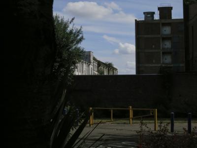 photograph “VIII.2015” par David Farreny — www.farreny.net — Royaume-Uni, United Kingdom, Grande-Bretagne, Great Britain, Angleterre, England, GB, UK, Londres, London, Greenwich, Woolwich, Charlton Riverside, bâtiments, buildings, immeubles, mur, wall, paroi, allée, alley, bitume, blacktop, asphalte, asphalt, fenêtres, windows, ciel, sky, nuages, clouds, plantes, plants, feuillage, foliage, barrière, métal, metal, fence, gate