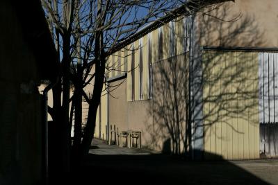 photograph “XII.2023” par David Farreny — www.farreny.net — France, Occitanie, Tarn-et-Garonne, Caussade, ville, city, bourgade, town, building, bâtiment, entrepôt, warehouse, métal, metal, tôle ondulée, corrugated iron, mur, wall, portail, gate, jaune, yellow, gouttière, drainpipe, arbres, trees, ombre, shadow, branches, branchages, tables, industrie, industry, angle, coin, corner, ciel, sky