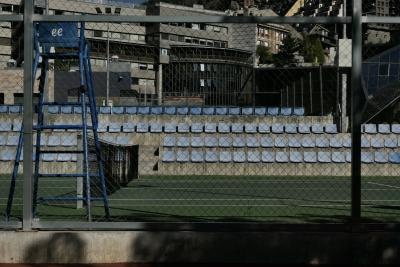 photograph “VIII.2023 — Joy cage” par David Farreny — www.farreny.net — Andorre, Andorra, Escaldes-Engordany, Prat del Roure, tennis, court, chaises, chairs, clôture, fence, grillage, fencing, métal, metal, filet, net, béton, concrete, immeubles, buildings, bâtiments, ville, city, chaise, arbitre, umpire, chair, fenêtres, windows