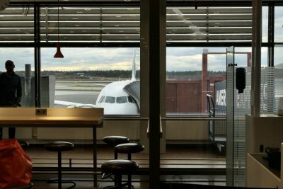 photograph “IV.2022 — Run away to the North, 299” par David Farreny — www.farreny.net — Suède, Sverige, Sweden, Uppland, Stockholm, Sigtuna, Arlanda, aéroport, airport, aérogare, terminal, avion, plane, fenêtres, windows, baie, vitre, verre, glass, lampe, lamp, passerelle, jet bridge, publicité, advertising, pub, ad, American Express, nuages, clouds, radiateurs, radiators, aile, wing, tabourets, stools, table, homme, man, voyageur, traveler, forêt, forest, horizon, voyage, travel, trip