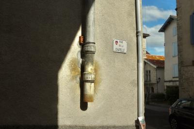 photograph “III.2025 — Whatever” par David Farreny — www.farreny.net — France, Occitanie, Tarn-et-Garonne, Caylus, village, maisons, houses, mur, wall, angle, coin, corner, plaque, plate, rue de la Chapelle, gouttières, drainpipes, métal, metal, ciment, cement, fenêtres, windows, volets, shutters, bois, wood, bleu ciel, sky blue, toits, roofs, tuiles, tiles, affiche, poster, voiture, car, câbles, cables, nuages, clouds, lampadaire, lamp, ruelle, alley