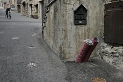 photograph “XI.2022 — Faces of home, 159” par David Farreny — www.farreny.net — Andorre, Andorra, Canillo, rue, street, bitume, blacktop, asphalte, asphalt, maisons, houses, pierre, stone, cycliste, cyclist, vélo, bike, portes, doors, fenêtres, windows, tuyaux, pipes, plastique, plastic, volets, shutters, bois, wood, escalier, staircase, marches, steps, rambarde, guardrail, voiture, car, symboles, symbols, féminin, female, signe, sign, Casa Farrenou, boîte à lettres, mailbox, métal, metal, domicile, home