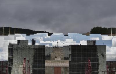 photograph “X.2023” par David Farreny — www.farreny.net — France, Occitanie, Pyrénées-Orientales, Font-Romeu-Odeillo-Via, Odeillo, four solaire, solar furnace, miroirs réfléchissants, reflecting mirrors, perspective, héliostats, heliostats, steerable mirrors, poteaux, poles, posts, béton, concrete, murets, low walls, pierre, stone, structures, métal, metal, colline, hill, arbres, trees, câbles, cables, fils, wires, nuages, clouds, ciel, sky, reflets, reflections, gris, grey, gray, toit, roof, tôle ondulée, corrugated iron, porte, door