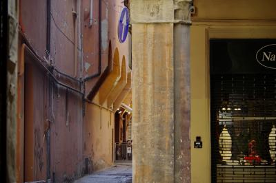 photograph “X.2019 — Lost in Bologna” par David Farreny — www.farreny.net — Italie, Italia, Italy, Émilie-Romagne, Emilia-Romagna, Bologne, Bologna, ville, city, murs, walls, croisement, carrefour, crossing, ciment, cement, coin, corner, angle, gouttières, drainpipes, tuyaux, pipes, métal, metal, magasin, shop, boutique, store, panneau, sign, flèche, arrow, câbles, cables, fils, wires, enseigne, Na, grille, railing, rideau, curtain, terrasse, terrace, café, lumières, lights, ruelle, alley